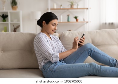 Young indian woman spend free time at home sit on cozy sofa at living room texting message in mobile app on smartphone. Millennial hindu lady chat in internet choose video to watch online using cell