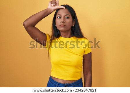 Young indian woman standing over yellow background making fun of people with fingers on forehead doing loser gesture mocking and insulting. 