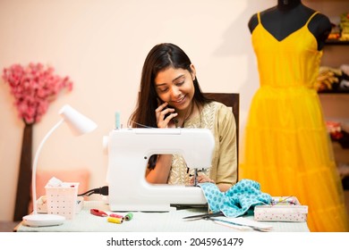 Young Indian Woman Tailor Or Fashion Designer Using Sewing Machine While Talking On  Smartphone In Garment Tailoring Workshop, Atelier Or Studio. Skill India. Small Business