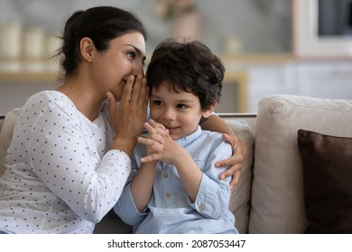 Young Indian woman telling secret whispering to interested little cute kid son, sharing confidential information or gossiping together at home, family enjoying trustful conversation.