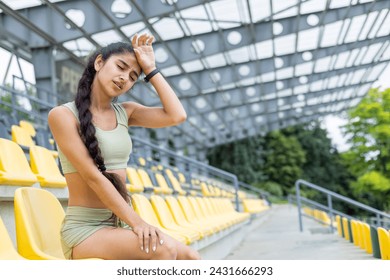 A young Indian woman in a tracksuit rests in a stadium, displaying fatigue after jogging. She wears a fitness smartwatch.