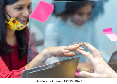 Young Indian Woman, Wearing Facemask Below Chin, Trying To Touch The Hand Of Her Colleague In Front Of Her, Plexiglass Barrier To Avoiding Direct Contact And Preventing Spreading Of Corona Virus