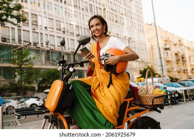 Young Indian Woman Wearing Sari Using Cellphone While Sitting On Scooter Outdoors