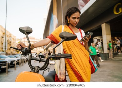 Young Indian Woman Wearing Sari Using Cellphone While Standing With Scooter Outdoors