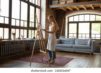Young indie artist woman working in studio  painting picture  standing at easel in creative loft space and artistic supplies  tools  drawing canvas  using paint paintbrush  Full length shot