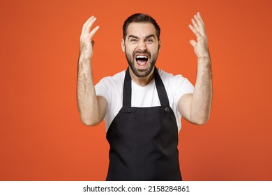 Young indignant angry stressed man barista bartender barman employee in black apron white t-shirt work in coffee spread hands scream shout isolated on orange background studio Small business startup
