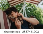 A young individual gently holding a snake while lying down outdoors. The scene captures a peaceful connection between human and reptile in a natural garden environment.