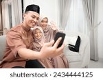 Young Indonesian family doing video call to other family and relatives with greetings gesture to the phone in happy during Eid mubarak in the living room at home. Islamic celebration concept.
