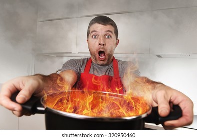 Young Inexperienced Home Cook With Apron Holding Pot Burning In Flames With Stress And Panic Face Expression In Fire In The Kitchen And Cooking Wrong Concept