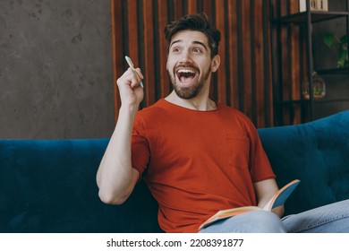 Young Insighted Man Wear Red T-shirt Writing In Notebook Point Finger Up With New Idea Sit On Blue Sofa Couch Stay At Home Hotel Flat Rest Relax Spend Free Spare Time In Living Room Indoors Grey Wall