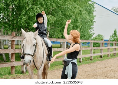 Young instructor guiding child in mastering horseback riding skills outdoors, with verdant trees and wooden fence creating serene backdrop for learning experience - Powered by Shutterstock