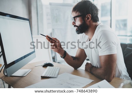 Young intelligent bearded man wearing eye glasses working at modern loft studio-office with desktop computer.Blurred background. Horizontal