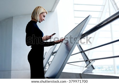 Young intelligent female phoning to partner via mobile phone while selecting information on high tech device, smart businesswoman holding cell telephone while exploring project on interactive monitor 