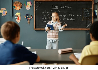 Young intelligent female student answering at blackboard holding notebook, while attentive classmates listening to presentation during lesson in classroom with creative kids crafts on blue wall - Powered by Shutterstock