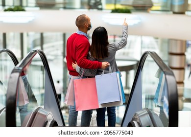 Young international couple in face masks standing on moving stairway, pointing at stores, shopping together in city mall during covid, back view. Seasonal sales and discounts, purchase and retail - Powered by Shutterstock