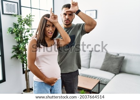 Young interracial couple expecting a baby, touching pregnant belly making fun of people with fingers on forehead doing loser gesture mocking and insulting. 