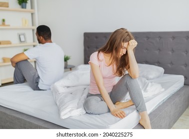 Young interracial couple having relationship difficulties, sitting on opposite sides of bed, not looking at each other at home. Black guy with wife going through marital crisis, having sexual problem