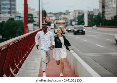 Young interracial couple holds hands, runs on bridge and laughs cheerfully on the background of the city and the road. The concept of love relationships and unity between different human races. - Powered by Shutterstock