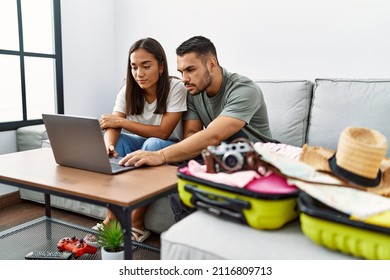 Young Interracial Couple Packing Summer Clothes In Suitcase Looking At Laptop Thinking Attitude And Sober Expression Looking Self Confident 
