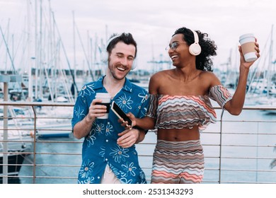 Young interracial couple is spending time together, listening to music on their headphones and drinking coffee. They are standing by a railing overlooking a harbor full of sailboats. Transgender woman - Powered by Shutterstock