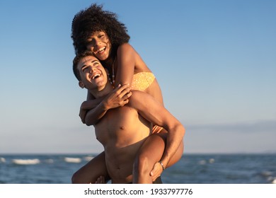 Young interracial couple in swimsuit having fun on the beach playing piggyback together in the summer.  - Powered by Shutterstock