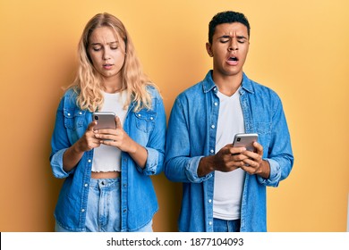 Young Interracial Couple Using Smartphone In Shock Face, Looking Skeptical And Sarcastic, Surprised With Open Mouth 