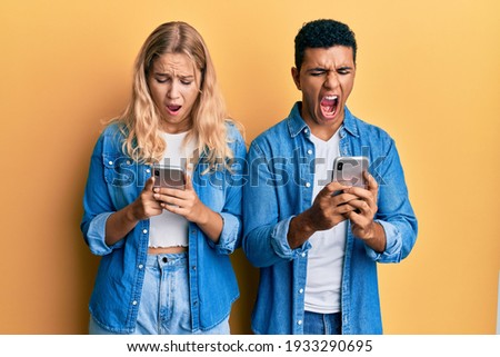 Young interracial couple using smartphone angry and mad screaming frustrated and furious, shouting with anger. rage and aggressive concept. 