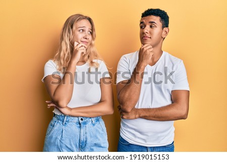 Young interracial couple wearing casual white tshirt with hand on chin thinking about question, pensive expression. smiling with thoughtful face. doubt concept. 