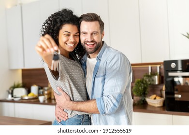 Young interracial married couple smiling cheerfully and showing keys from a new apartment, hugging and looking at camera, standing in the modern kitchen of a new home, real estate and family concept - Shutterstock ID 1912555519
