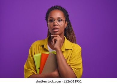 Young intrigued pretty African American woman student holds workbooks looks at camera with frightened face, opening mouth in surprise or fear stands on purple background. Education, training, exams