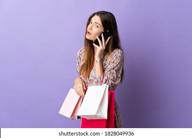 Young Ireland Woman Isolated On Purple Background Holding Shopping Bags And Calling A Friend With Her Cell Phone