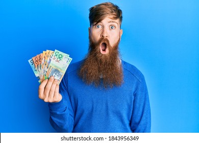 Young irish redhead man holding australian dollars scared and amazed with open mouth for surprise, disbelief face 