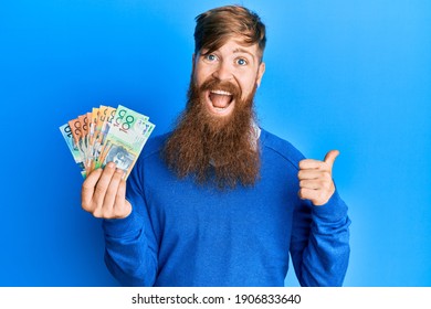 Young irish redhead man holding australian dollars pointing thumb up to the side smiling happy with open mouth 