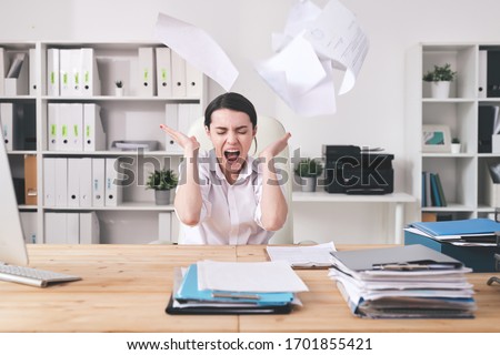 Young irritated or tired female office manager throwing financial papers over her head while expressing fury during work with documents