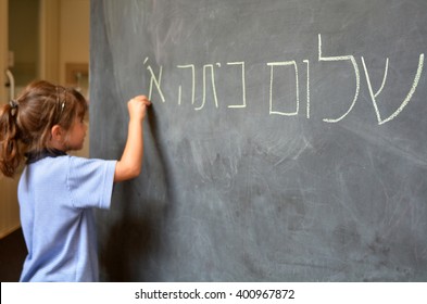 Young Israeli Jewish Girl (Age 5-6) Writes Hello First Grade Greetings In Hebrew (Shalom Kita Alef) On Chalkboard In Israeli Primary School At The Beginning Of The School Year. Real People.Copy Space