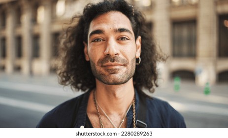 Young italian guy with long curly hair and stubble stands on the sidewalk next to the road, looks around and smiles - Powered by Shutterstock