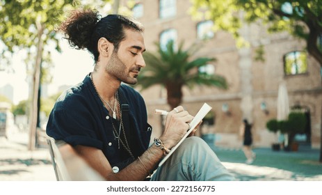 Young italian guy with ponytail and stubble sits on street bench and makes sketches with pen on piece of paper - Powered by Shutterstock