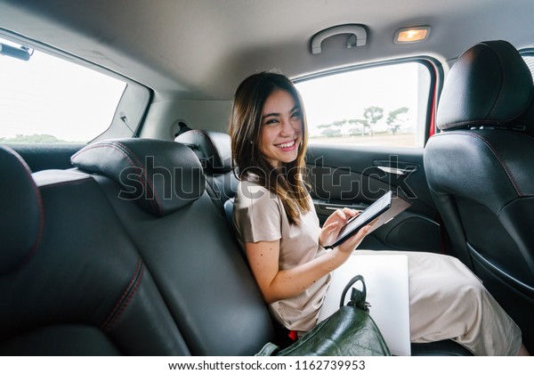 A young Japanese Asian woman sits and reads\
her e reader while sitting in the backseat of a car she booked via\
a ride hailing app. She is smiling happily as she reads and is\
driven to her destination.