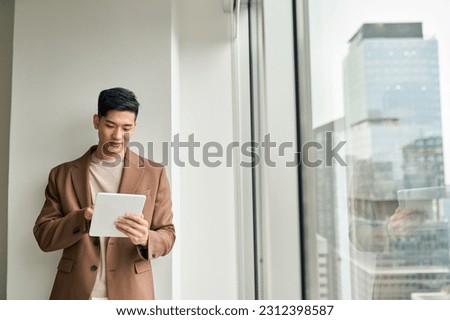 Young Japanese business man, Asian company manager or entrepreneur wearing suit holding digital tablet pc standing near big office window checking financial services or corporate software technology.
