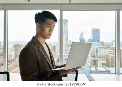 Young Japanese businessman employee holding laptop standing in big office. Busy professional Asian business man executive leader working on laptop using computer technology in corporate space. - Powered by Shutterstock