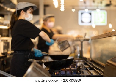 Young Japanese Female Chef Cooking With A Wok In An Open Kitchen. She Has A Pony Tail, Wears A Hat, Gloves And Mask.