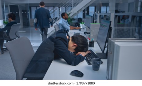 Young Japanese Manager Is Tired At His Work Place With Desktop Computer And Falls Asleep. Diverse Multi-ethnic Business People Work On Computers In Modern Open Office.