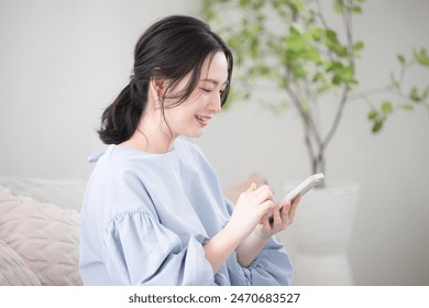 A young Japanese woman with her mobile phone, thinking in her beautiful living room. She looks at the display and taps to operate it. - Powered by Shutterstock