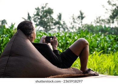 Young Javanese man lying on brown bean bag while playing mobile games or chatting someone. Relaxed Indonesian people enjoying sunset at garden and watching his smartphone or mobile cell phone. - Powered by Shutterstock