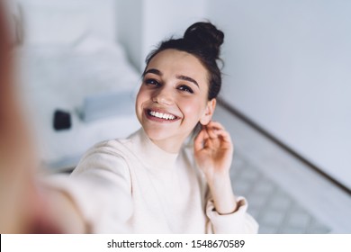 Young jolly female in light soft sweater holding mobile phone at bedroom with hand behind head and taking happy selfie 