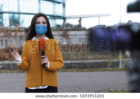 Young journalist with medical mask and microphone working on city street. Virus protection