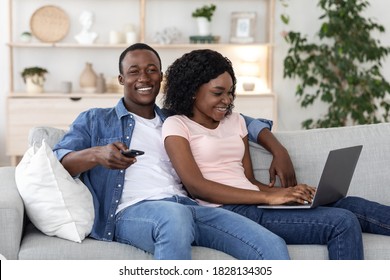 Young joyful african american couple watching TV and using laptop, spending weekend together at home, copy space. Relaxed black woman using laptop, happy man holding TV remote, living room interior