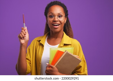 Young joyful African American woman student with enthusiastic smile raises pen and holds books to study for exam wants to enter college or university stands in purple studio. Back to school concept - Shutterstock ID 2200029291
