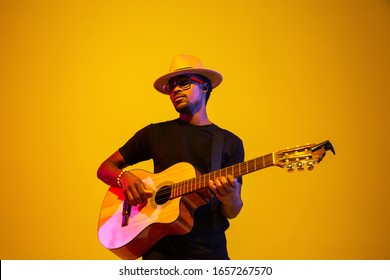 Young and joyful african-american musician playing guitar and singing on gradient orange-yellow studio background in neon light. Concept of music, hobby, festival. Colorful portrait of modern artist. - Powered by Shutterstock