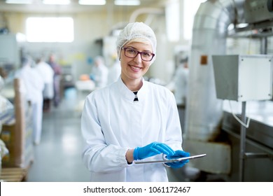 Young joyful beautiful female worker in sterile cloths holding a tablet and smiling for the camera near factory production line. - Shutterstock ID 1061172467